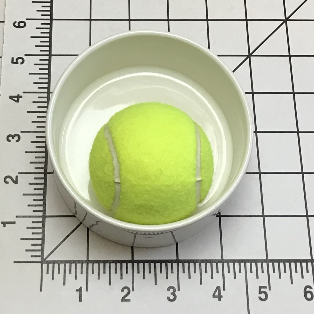 Tennis ball in a tiny white cup on a grid background with a ruler for scale.