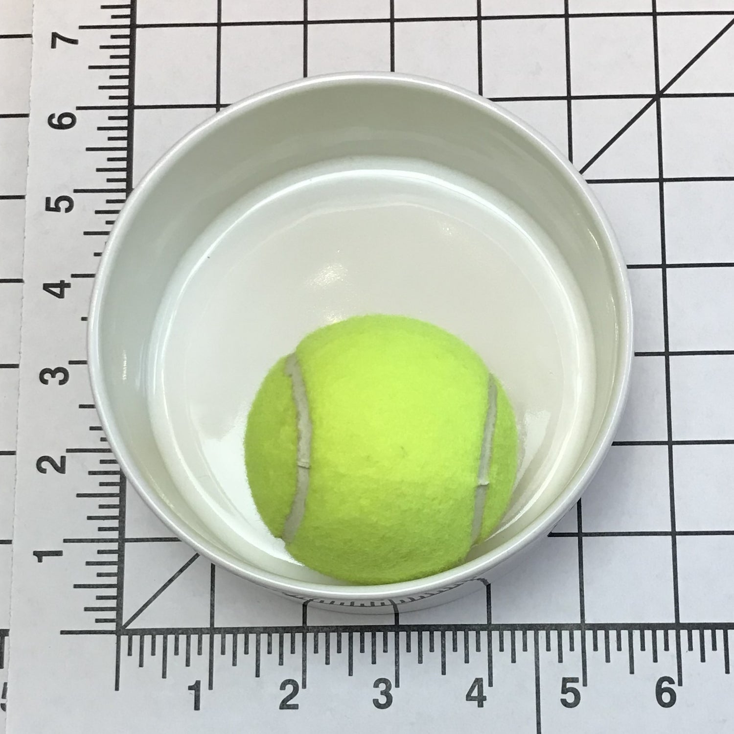 Tennis ball in a small size white bowl on a grid background with a ruler for scale.