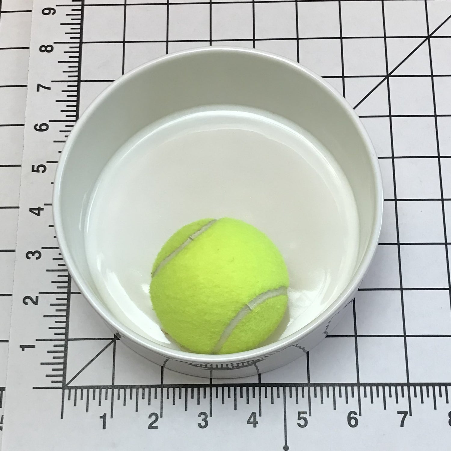 Yellow tennis ball in a medium size white bowl on a grid background with a ruler.