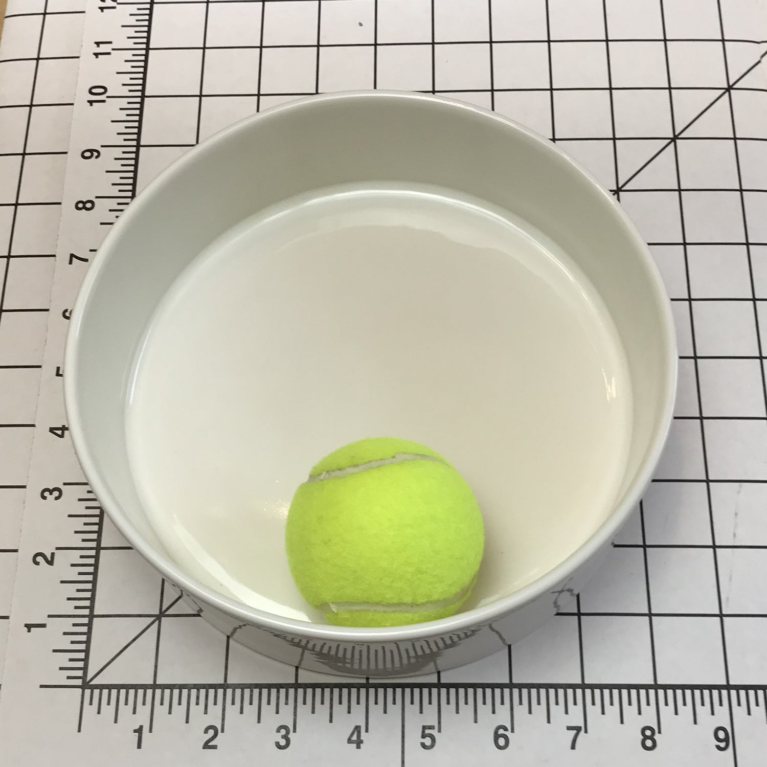 Tennis ball inside a large white bowl on a grid background with a ruler.